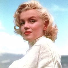 Marilyn Monroe wearing a sweater and lipstick and her classic blond hairdo. Posing for the camera.