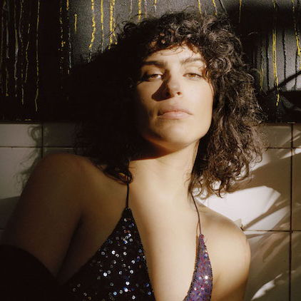 Leila laying against a wall wearing a dress and looking intently at the camera. There is light shining on her from a window.