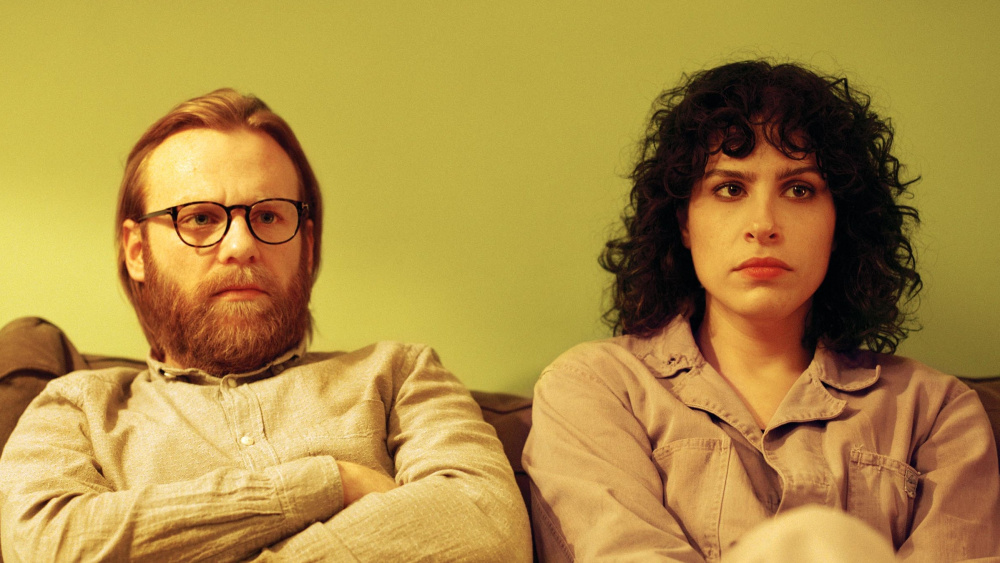 Gabe and Leila sitting on a couch both with serious expressions and arms crossed.