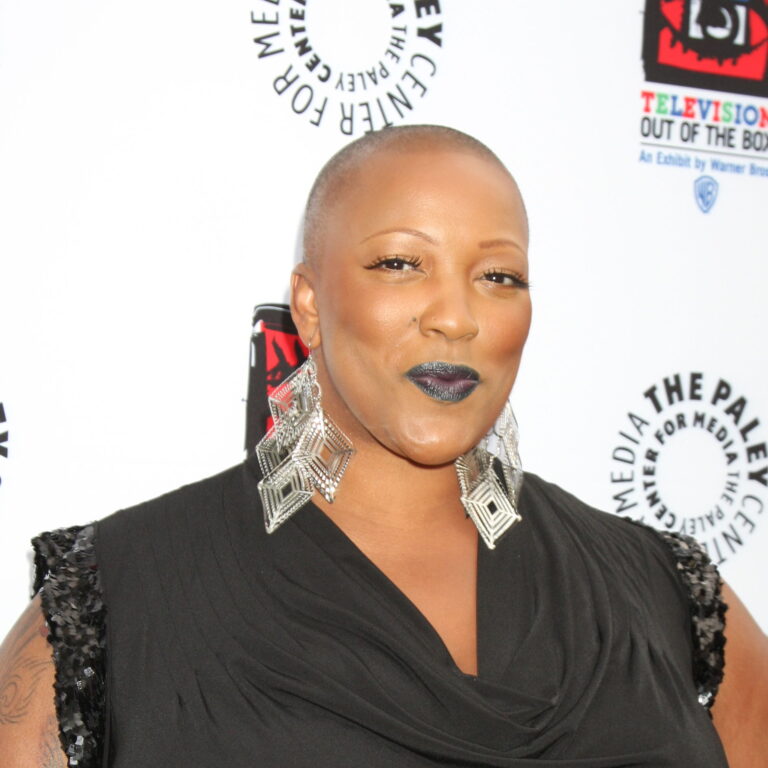 Frenchie Davis at a carpet event wearing large earrings and a dress, smiling.