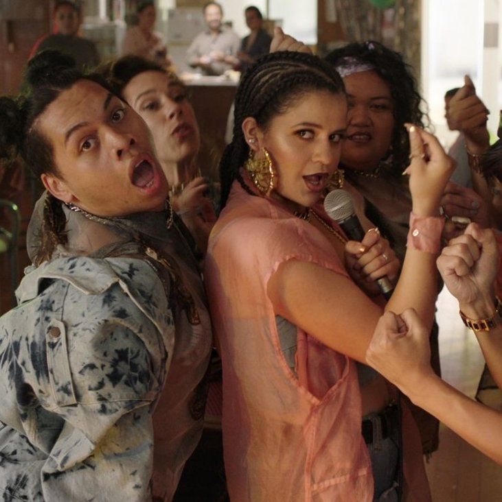 Image of several of the cast members who are POC, looking at the camera while they are dancing together during the day inside a building. Everyone has on fashionable clothes.