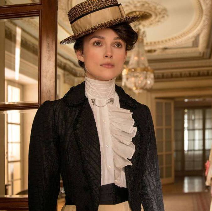 Colette dressed in the clothing from the time, walking and looking straight ahead inside a beautiful house.
