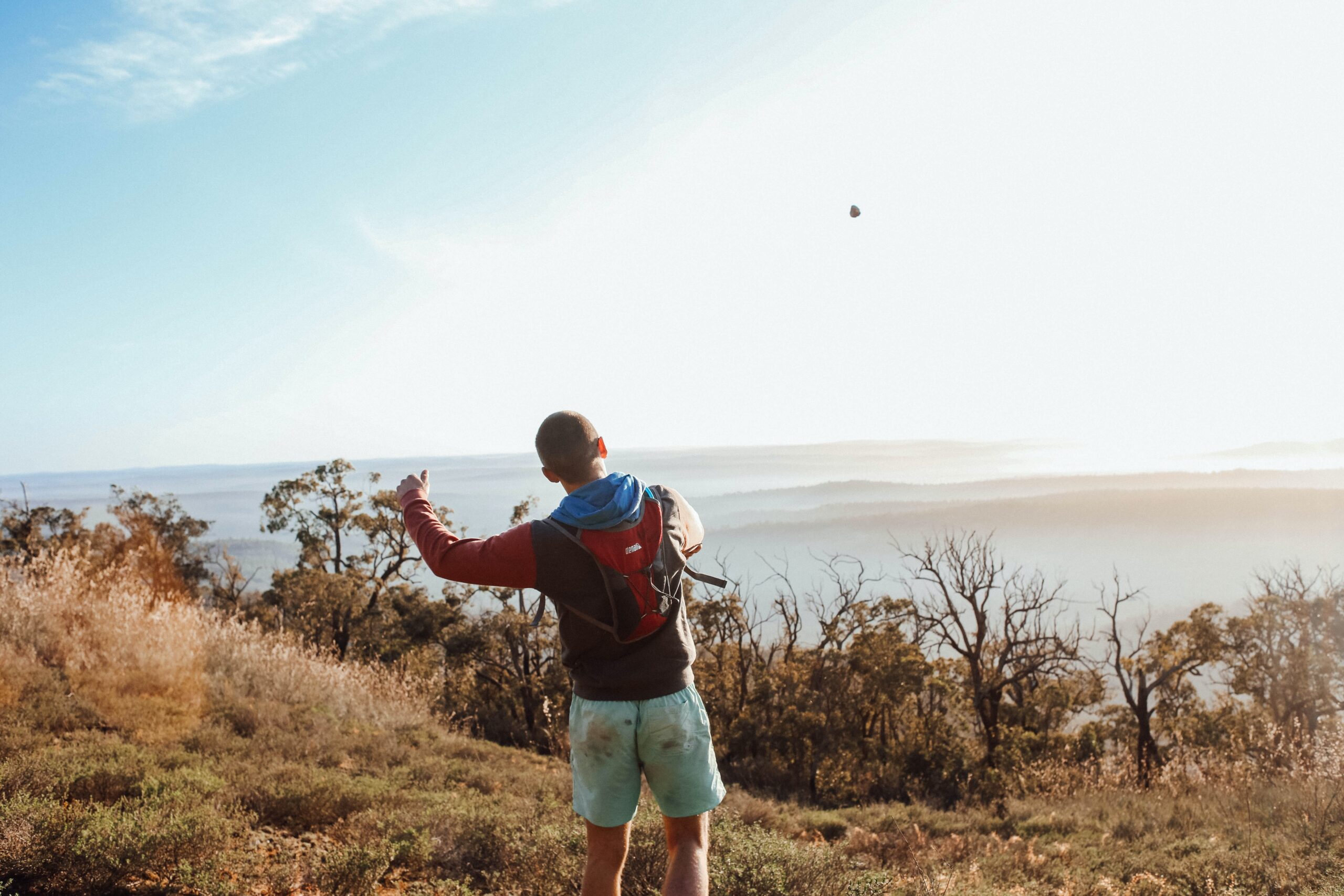A man is turned looking at the ocean from a hill throwing a pebble in the air.