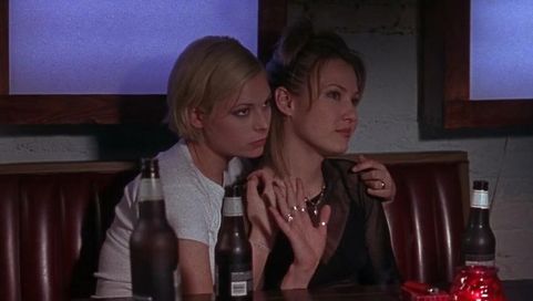 Alyssa and her girlfriend sitting at a booth together with beers nearby. Alyssa is waving to someone off camera while her girlfriend is holding her.