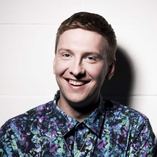 Joe Lycett con una colorida camiseta verde azulado y sonriendo a la cámara. Está contra un fondo de ladrillo blanco.
