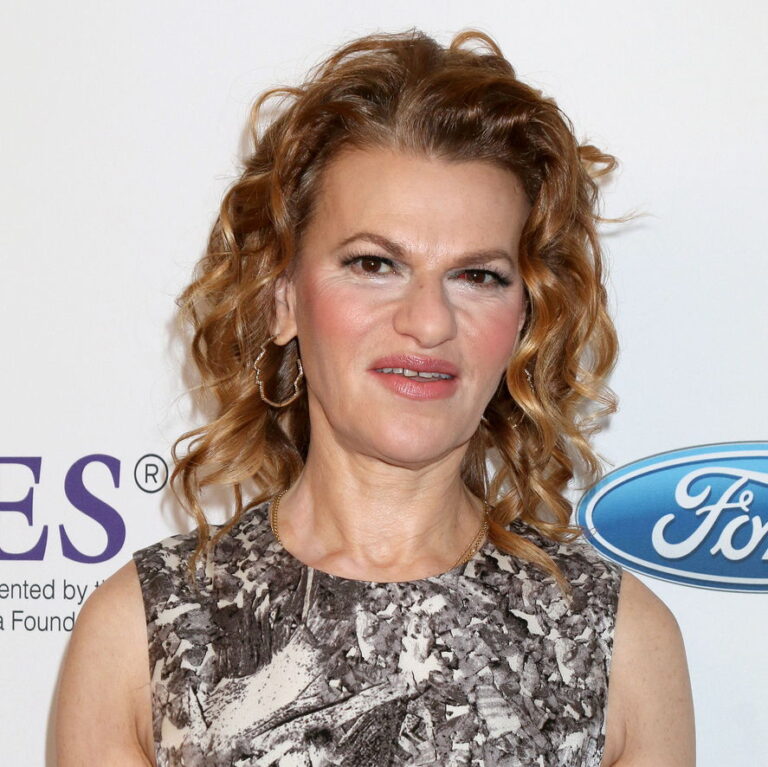 Sandra Bernhard waring a tan dress, on a stage with a white background. She is looking at the camera smiling slightly.