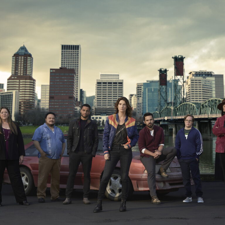 Characters of Stumptown posing in front of a car with the city behind them.