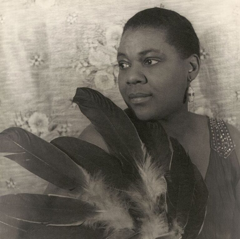 Black and white image of Bessie, posing with large feathers looking to her side, wearing a dress.