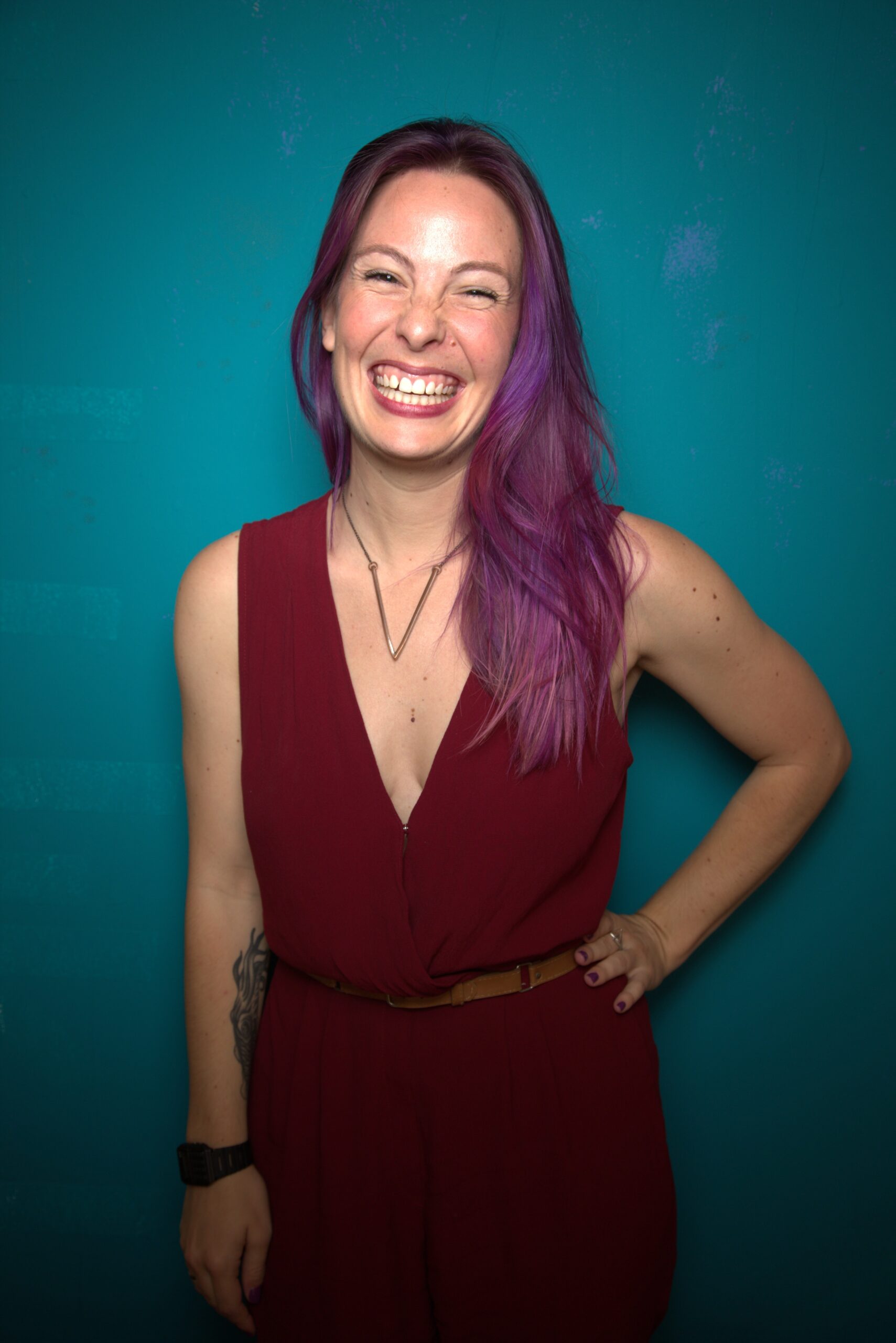 Photo of Amber Rollo wearing a red dress, purple hair and a big smile, against a teal background.