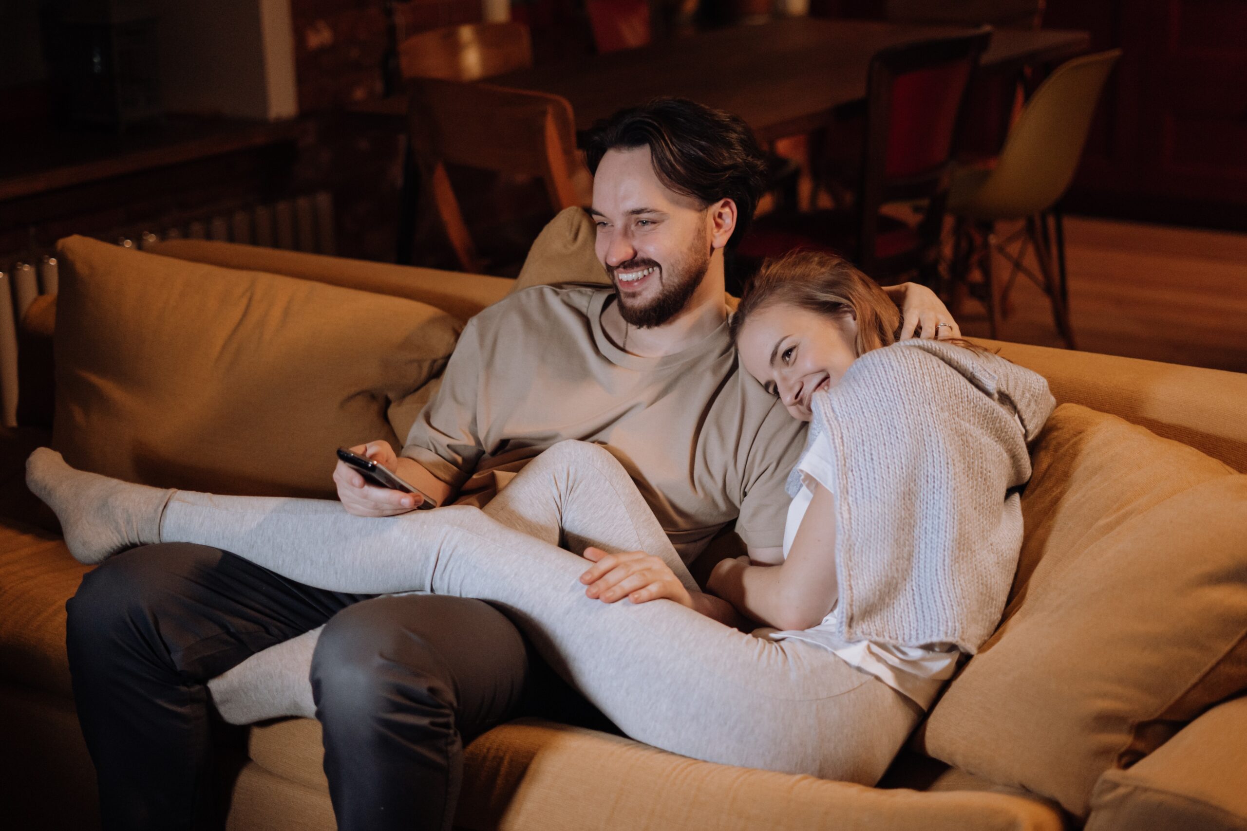A young attractive white couple laying together, the guy is smiling with the remote and the girl has her feet up smiling also.