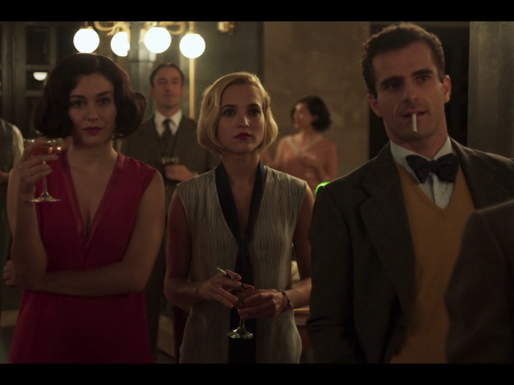 Carlota standing between a man and woman all looking intently foward at a party in a large house.