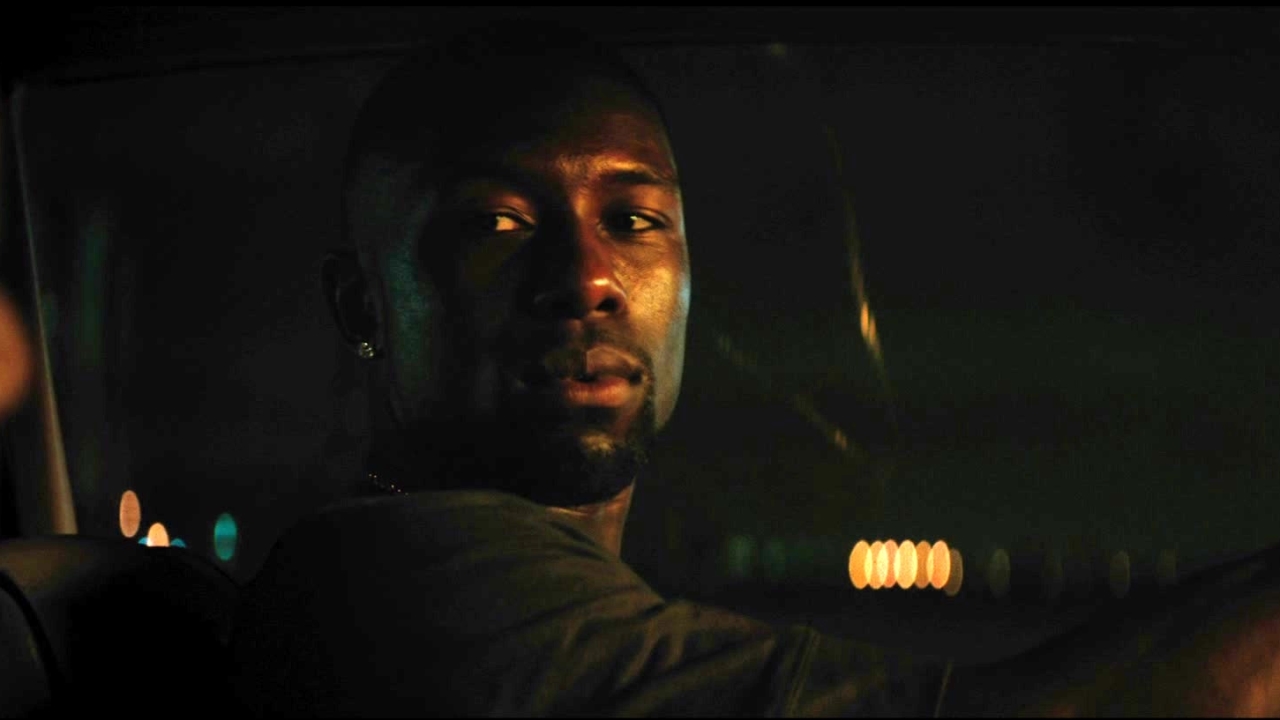 Chiron sitting in his car with hands on the wheel looking to his side at someone.