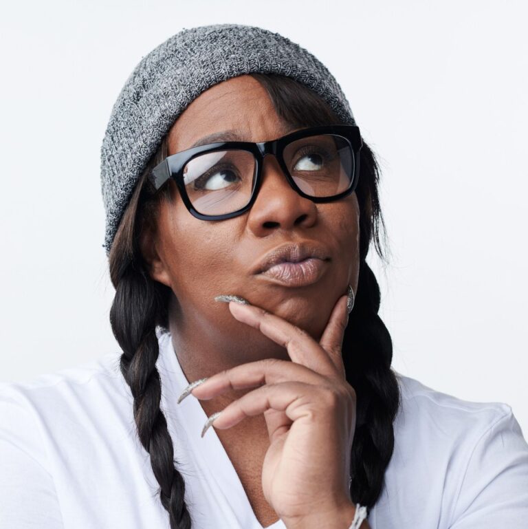 Franqi French llevándose la mano a la barbilla mirando pensativa hacia el techo. Lleva el pelo recogido en trenzas con un gorro y gafas.