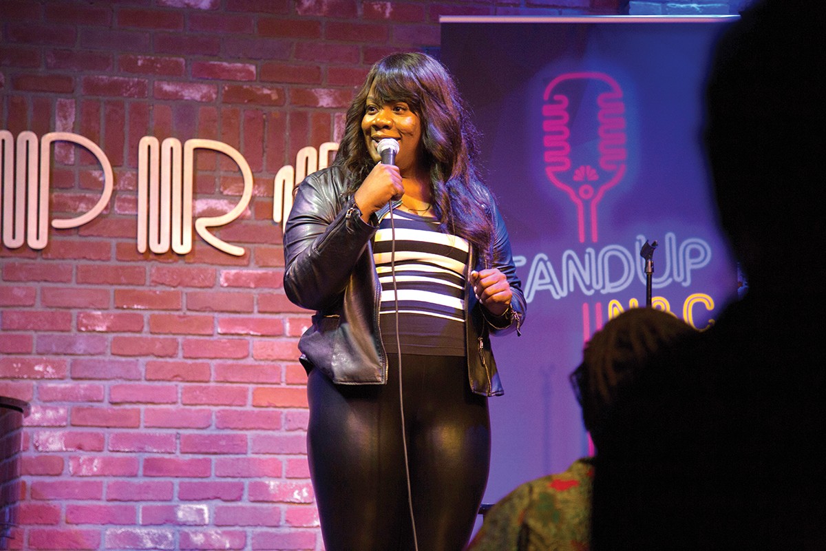 Franqi French doing a standup routine, she is holding a microphone smiling, onstage.