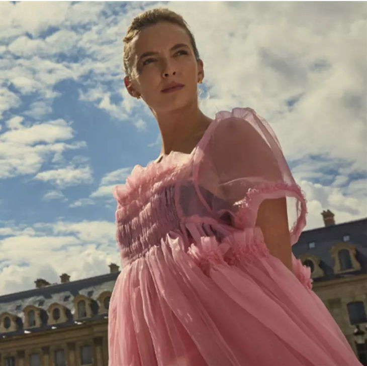 Vianelle wearing a pink and light dress with her hands behind her back looking to the distance with her eyes set on something.