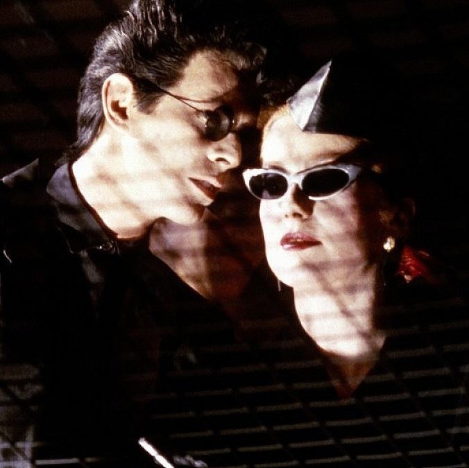 Bowie talking to Catherine at an event. Both are wearing stylish black clothes and sunglasses.