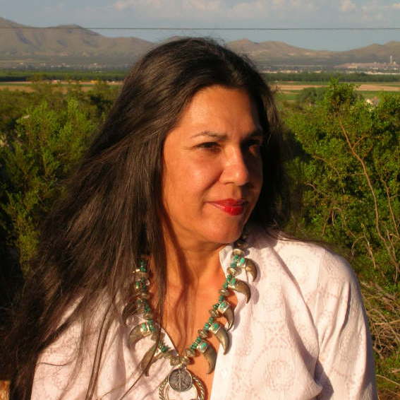 A portrait of Ana Castillo smiling facing the sun at an outdoor area.