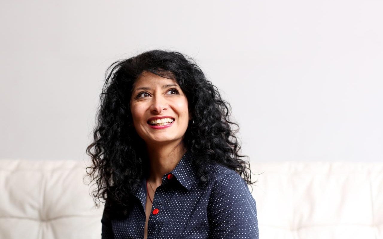 Shappi Khorsandi smiling and looking at a distance. She is sitting on a large couch wearing a buttoned up shirt and has wavy curly hair.