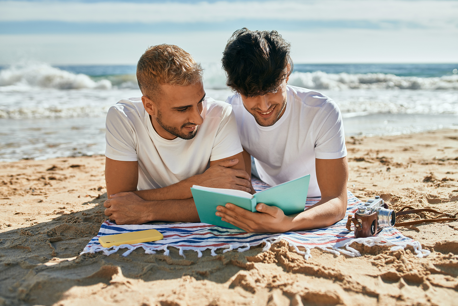 Dos chicos sobre una manta en la playa leyendo un libro.
