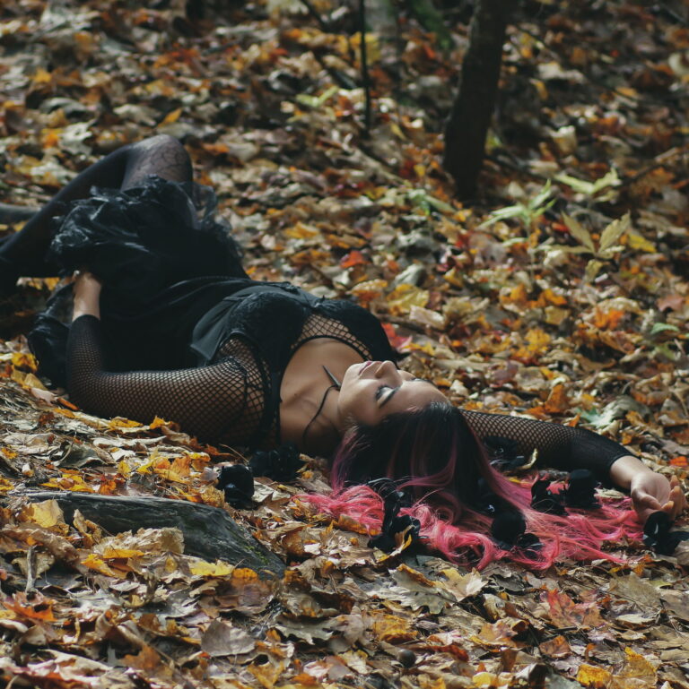 A woman with dyed hair and black dress lays on the ground covered in leaves.