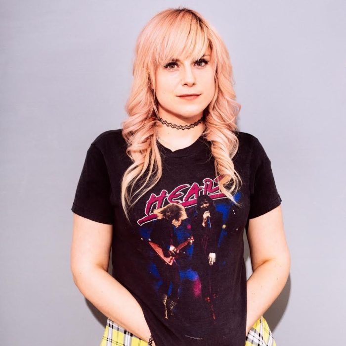 Lane More with dyed hair and a rocker shirt smiling with her hands in front.