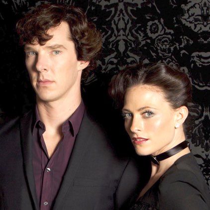 Sherlock and Irene standing next to eachother giving a look to the camera in front of a black wall.