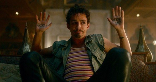 Klaus sitting with his legs crossed and his arms up. On his palms you can see a Hello and Goodbye tattoos