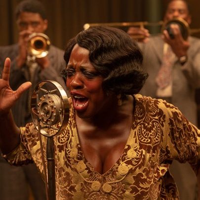 Ma Rainey, wearing a gold dress, singing passionately onto a mic with her trumpet band members.