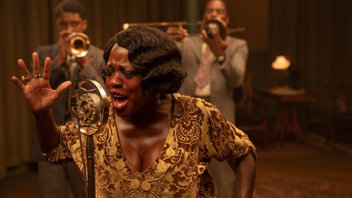 Ma Rainey, con un vestido dorado, cantando apasionadamente en un micr&oacute;fono con los miembros de su banda de trompeta detr&aacute;s de ella.