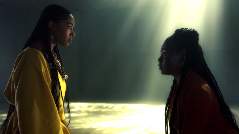 Bilquis kneeling in front of another god from the series. There only light visible is from an opening in the ceiling.