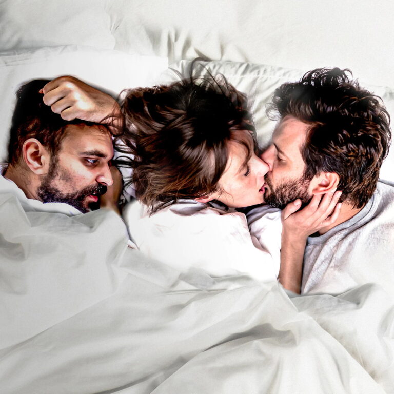 Zoe, Lucks, and Tom in bed together under the covers. Zoe is kissing Tom facing him while Lucks faces them.