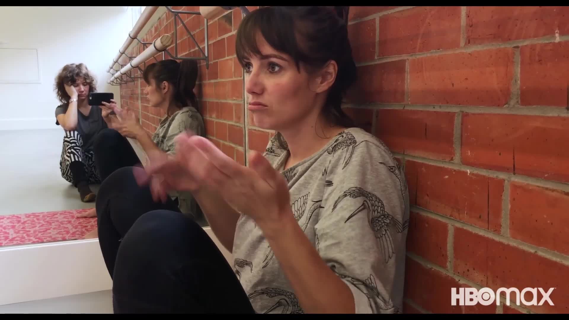 Zoe hablando sola sentada contra una pared de ladrillos mientras una mujer se ve en el espejo junto a ella, grabandola.