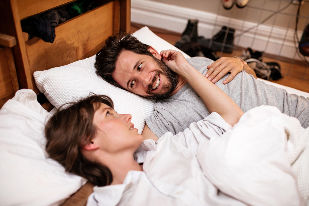 Zoe and Tom laying in bed together. Zoe has her hand on his face while he smiles at her.