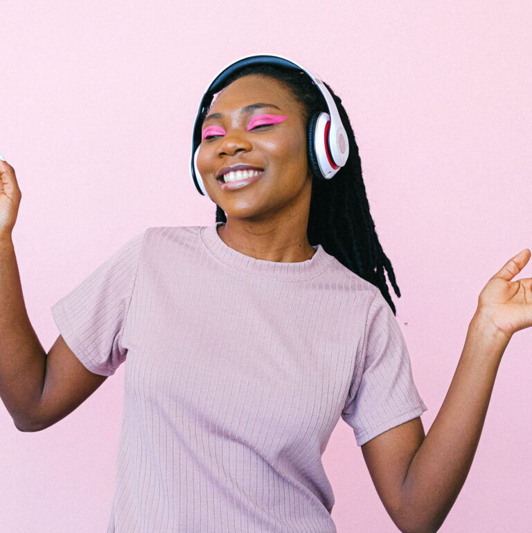 Image of black woman with headphones on and her eyes closed, dancing by herself. There is a purple/pink background.