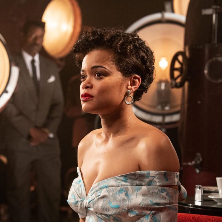Image of Lady Day, sitting with cameras and lights all around her, she is looking at a distance and has a low cut dress.