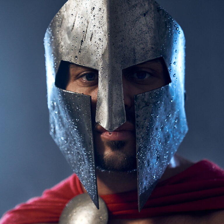 Portrait of spartan warrior standing and looking at camera. Close up of mature man in red cloak and helmet with water drops posing in dark atmosphere.