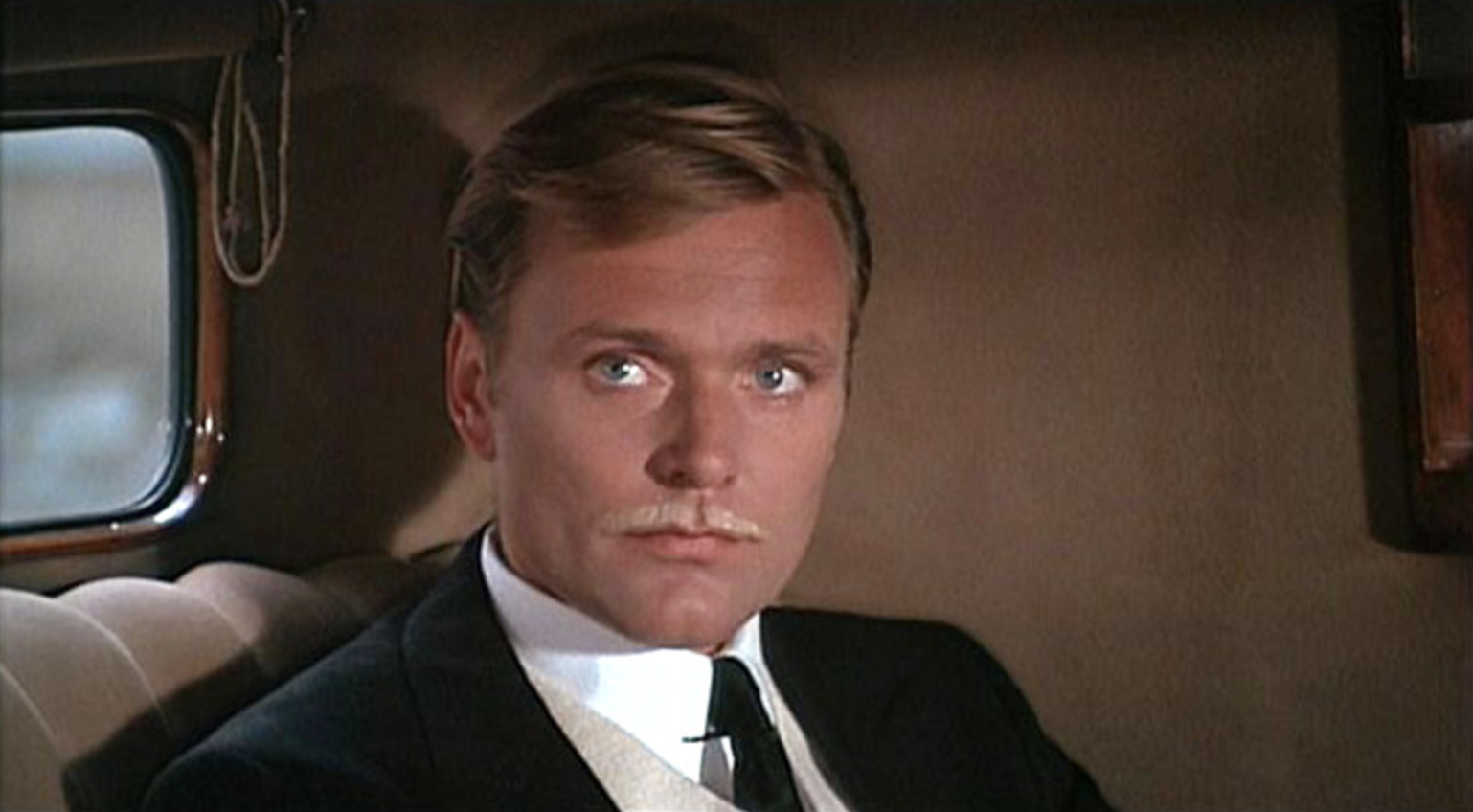 Baron Maximilan wearing a black suit and tie, sitting in a car looking at someone off camera.