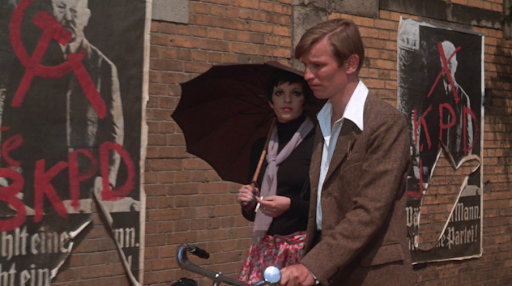 Brian walking with sally down a street with large posters behind. Its a sunny day.