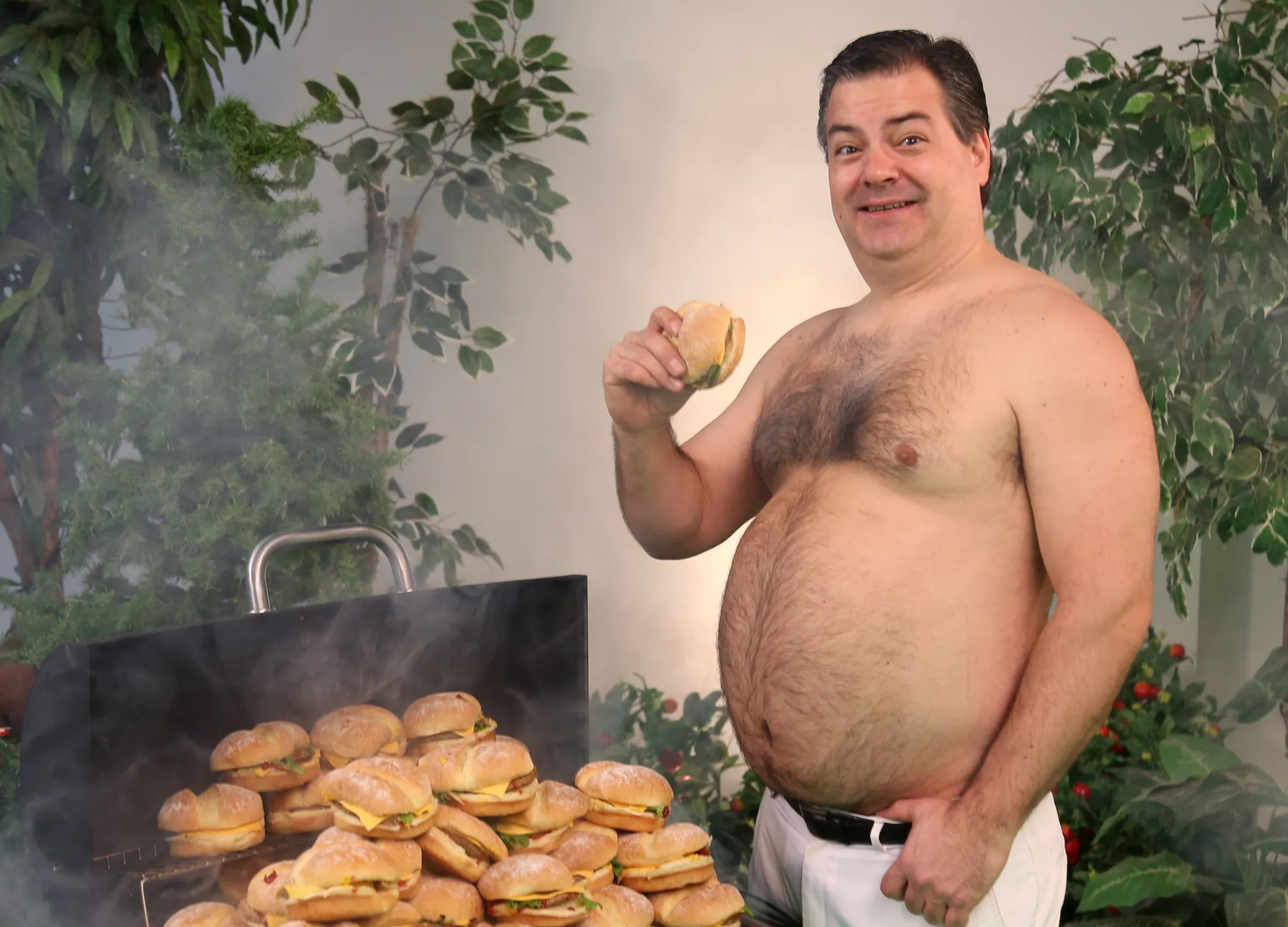 Randy with no shirt smiling while holding a cheeseburger next to a grill covered in a mountain of cheeseburgers
