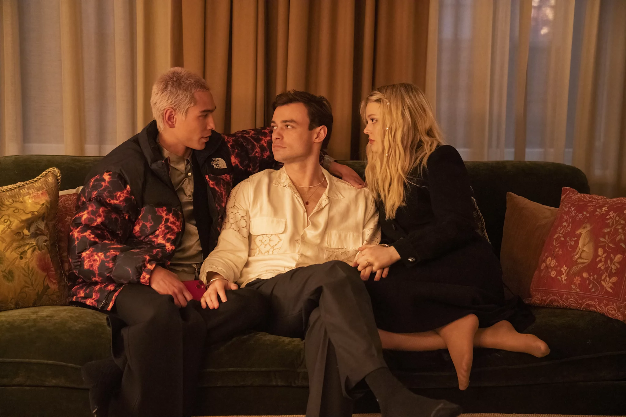 Gossip Girl characters Aki, Max, and Audrey sitting on a couch holding hands and looking serious