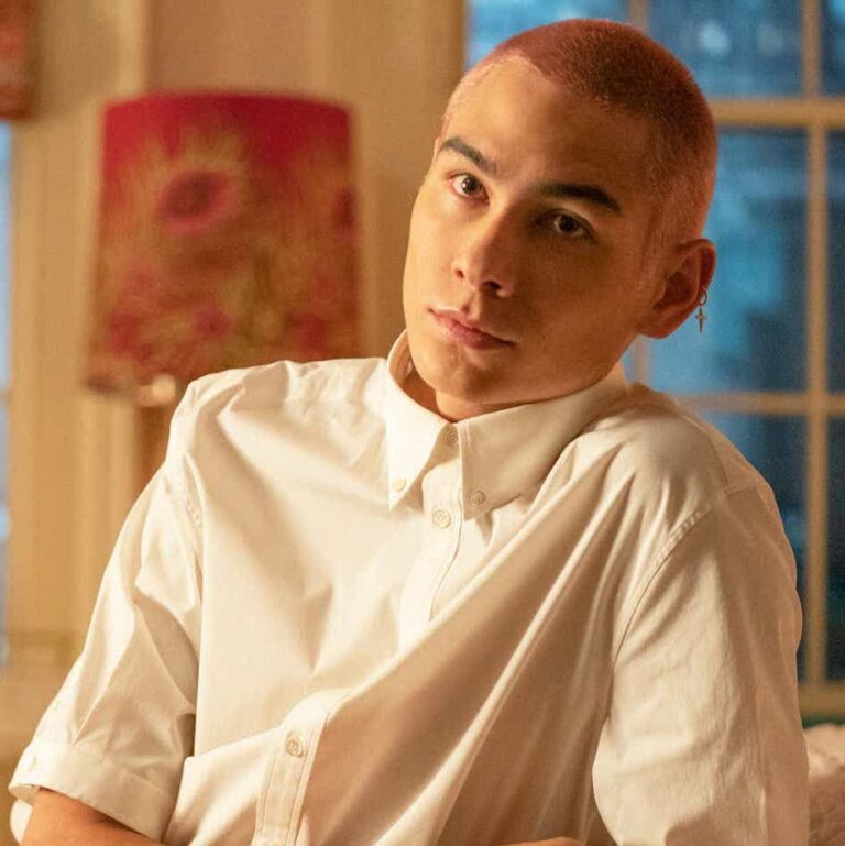 Aki leaning on his side looking at someone off camera. He has short buzzed dyed hair.