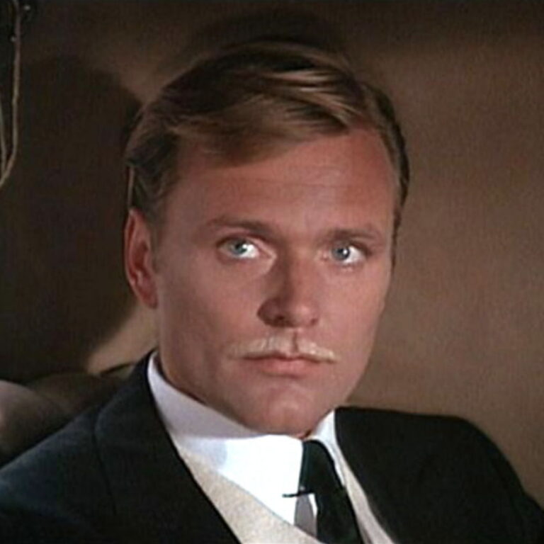 Baron Maximilan wearing a black suit and tie, sitting in a car looking at someone off camera.