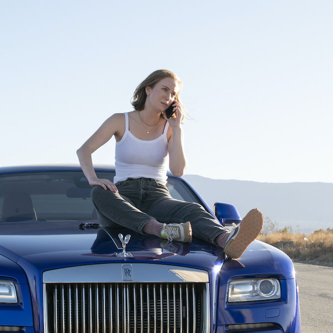 Ava wearing a white tanktop and jeans, sitting on top of sleek blue car, and is on the phone with a concerned look.