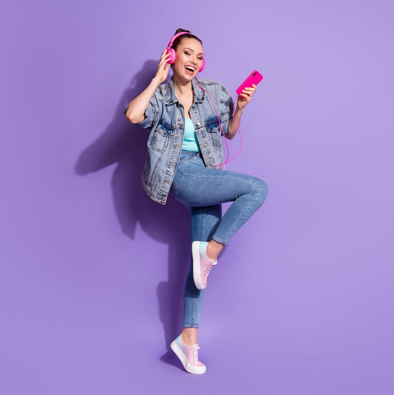 An attractive woman wearing pink headphones against a purple background, jumping in the air and smiling.
