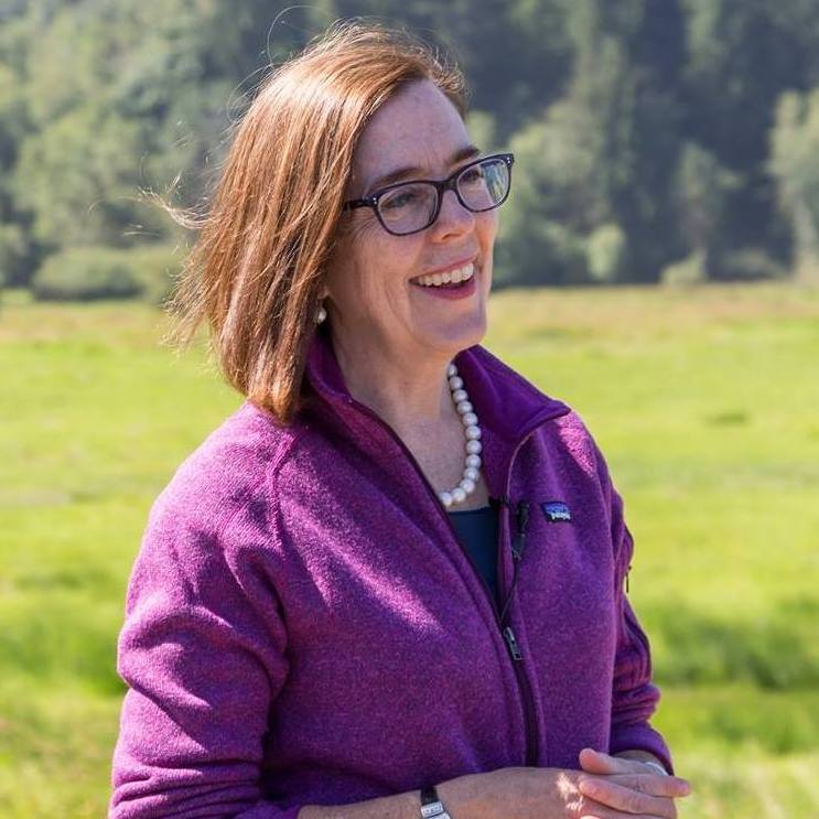 Kate Brown smiling holding her hands together in a local field, wearing purple representing.