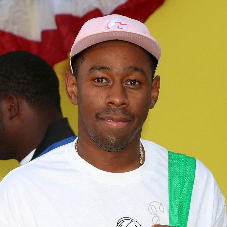 Tyler at a carpet event smiling wearing a cap t shirt and a backpack.