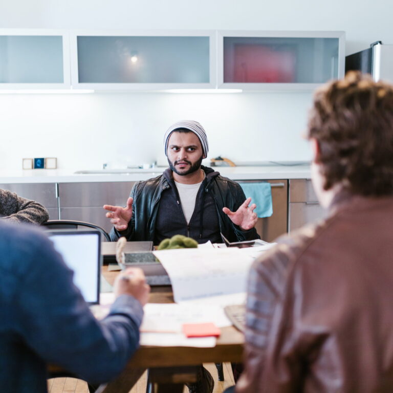 A multi ethnic man wearing a beanie and jacket talks to two friends who are doubtful of what he has to say.