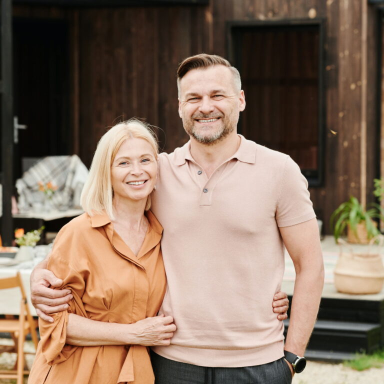 An older white couple smiles while outside. The husband has his hand around her waist.
