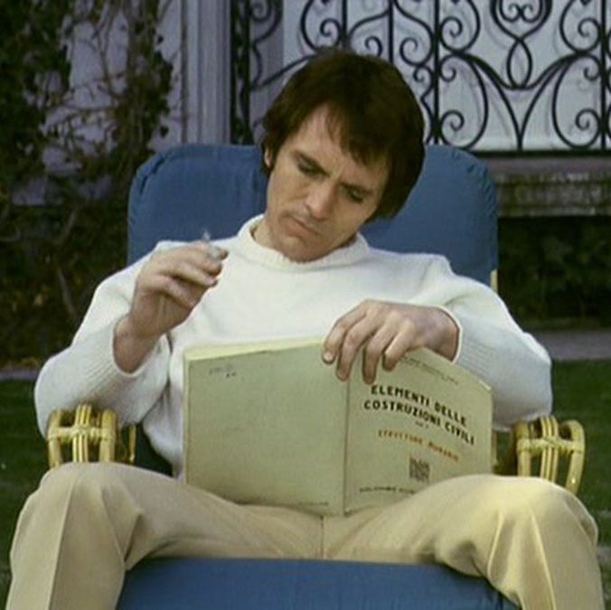 The Visitor sitting with his legs spread reading a book and smoking a cigarette outside.