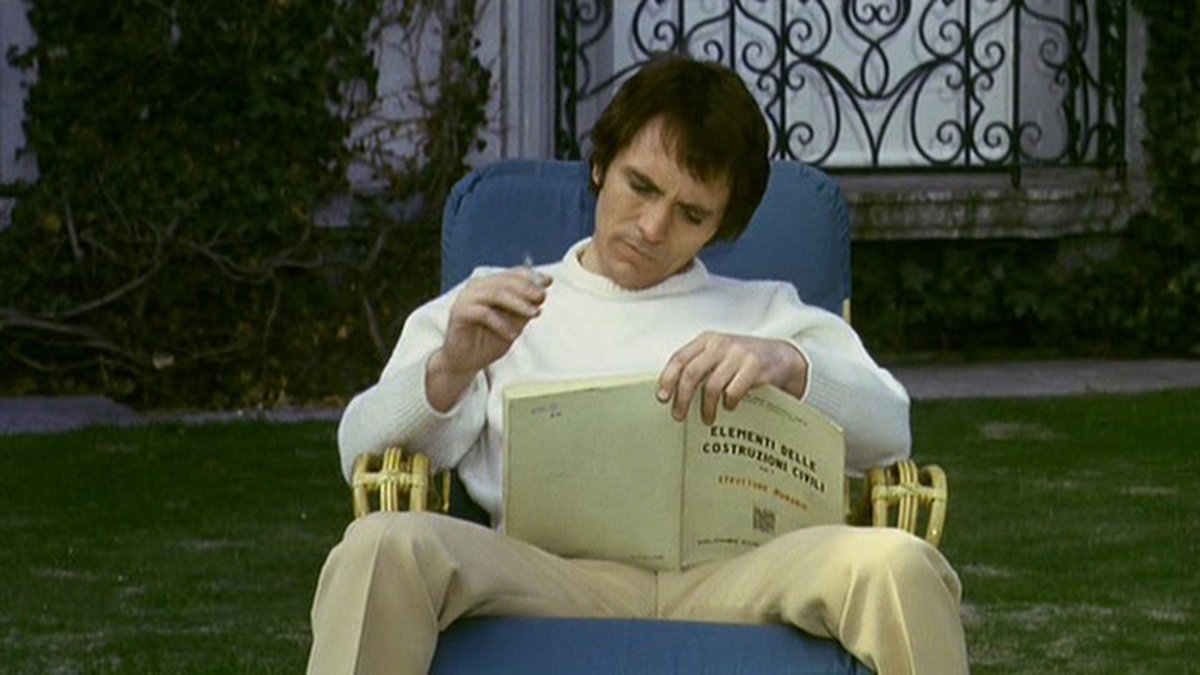 The Visitor sitting with his legs spread reading a book and smoking a cigarette outside.